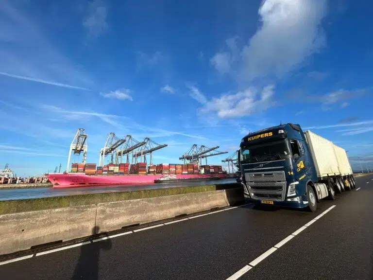 A blue and yellow container truck drives past an international container ship. On the truck are two white containers perfect for international transport.