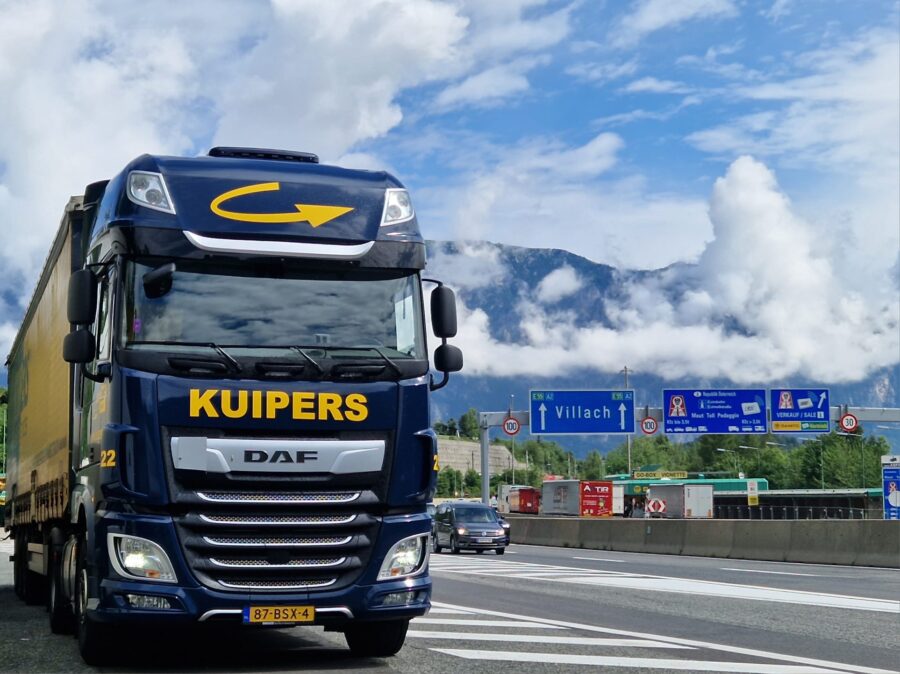 A blue and yellow Kuipers Logistics truck is seen passing the border crossing near Villach. "Villach" is visible on the traffic sign behind the truck.