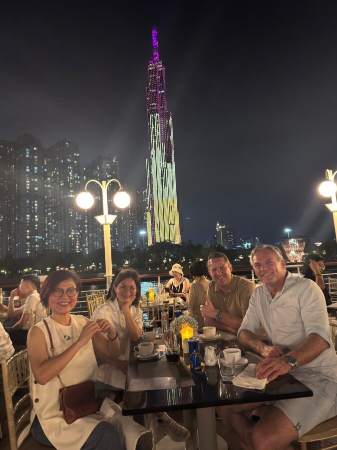 Wouter and Jos sit at a table with two women. The women sit on one side, and Wouter and Jos sit on the other. Even though it’s already dark, everyone is still wearing summer clothes. In the background, a large, tall, lit building can be seen.