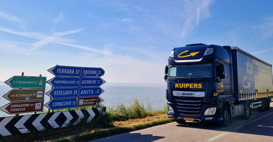 A Kuipers Logistics truck is driving on a road between Ravenna and Ferrara. It passes road signs with directions to four cities to the left: Ferrara, Portomaggiore, Ostellato, and Comacchio. And three cities to the right: Ravenna, Alfonsine, and Anita.