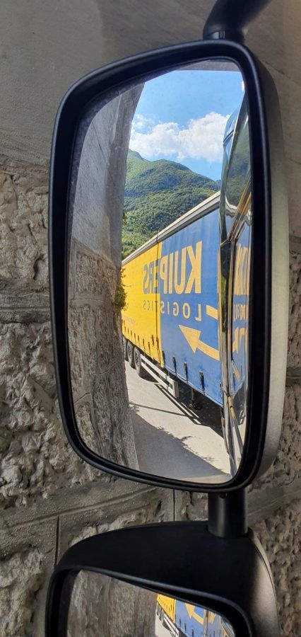 A Kuipers Logistics trailer is visible in the side mirror of a truck. The trailer is blue and yellow, and the side mirror has a tall rectangular shape.