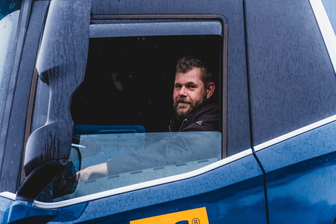 ingezoomde foto van een vrachtwagenchauffeur met baard die uit zijn open raam kijkt. De vrachtwagen is blauw. En heeft zwarte spiegels. Verder is de vrachtwagen nat van de regen.