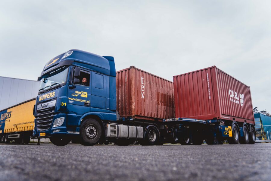 Kale containerchauffeur zet twee, rode containers naast elkaar. Hij rijdt in een blauwe DAF XF440 met een broshuis containerchassis. Voorbereiding op LCL transport.