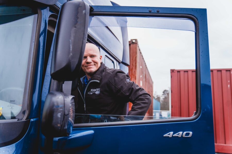 Kale vrachtwagenchauffeur stapt in de vrachtwagen. De vrachtwagen is iets nat van de regen. Het raam van de deur is open en de chauffeur kijkt lachend door het raam naar iets in de verte.