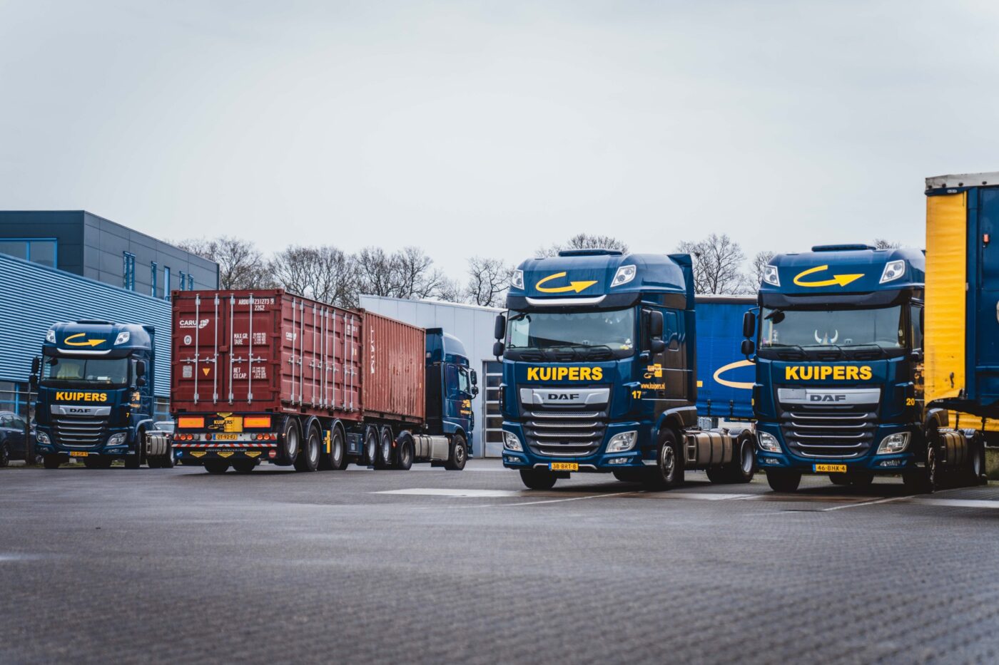 De containerchassis van Kuipers Logistics rijdt van de parking. Links van de chasssis is nog één vrachtwagen te zien. Rechts zijn nog twee vrachtwagens ze zien, met daarnaast en achter nog een trailer.