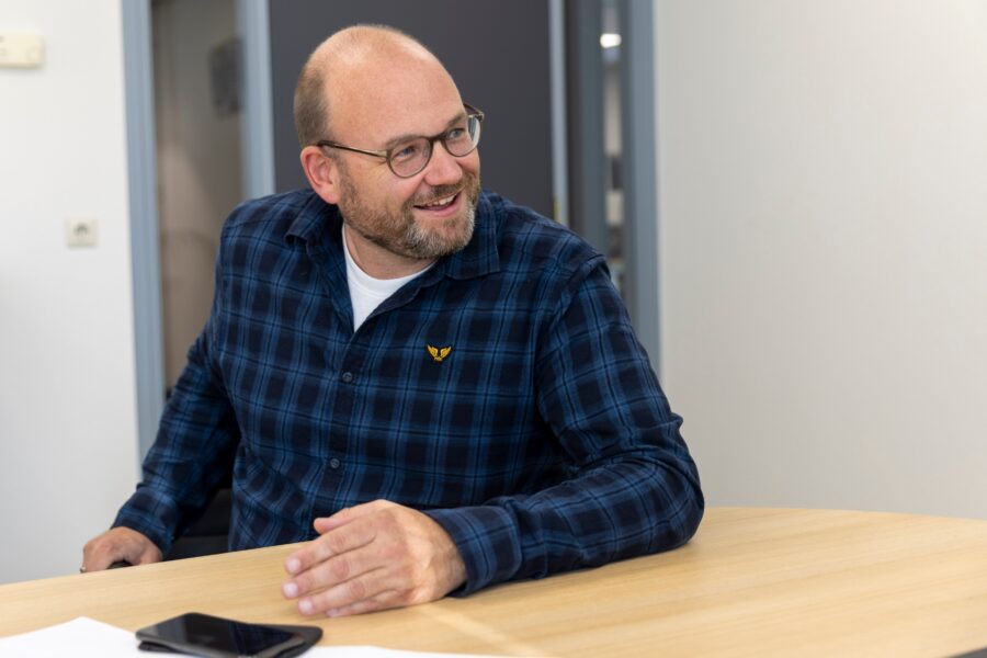 Philip Vollenbroek zit voor zijn interview schuin aan tafel. Hij is enthousiast aan het praten en heeft zijn linker arm op tafel en met zijn rechter arm houd hij de leuning van zijn stoel vast. Hij heeft een blauwe geblokte blouse aan met daaronder een wit T-shirt.