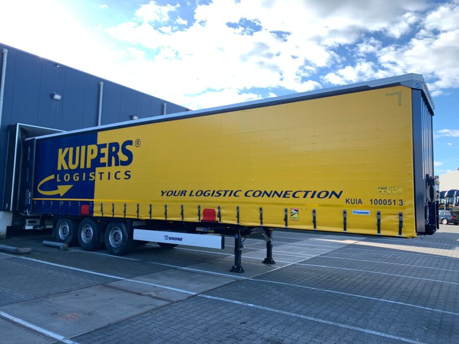 Op de foto is de linker kant van een nieuwe KRONE Profi Liner SDP 27 eLHB50-CS van Kuipers Logistics te zien. de trailer staat aan het dok en is van een geel met blauwe opdruk voorzien.
