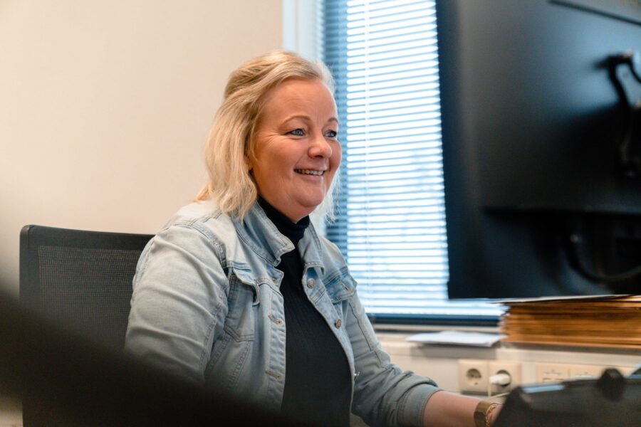 Jacqueline Mulder zit lachend achter haar bureau bij kuipers logistics. Ze heeft een zwarte coltrui aan met daar overheen een lcihtblauw spijkerjasje. Ze heeft blond haar dat tot de schouders komt en half omhoog zit en blauwe ogen.