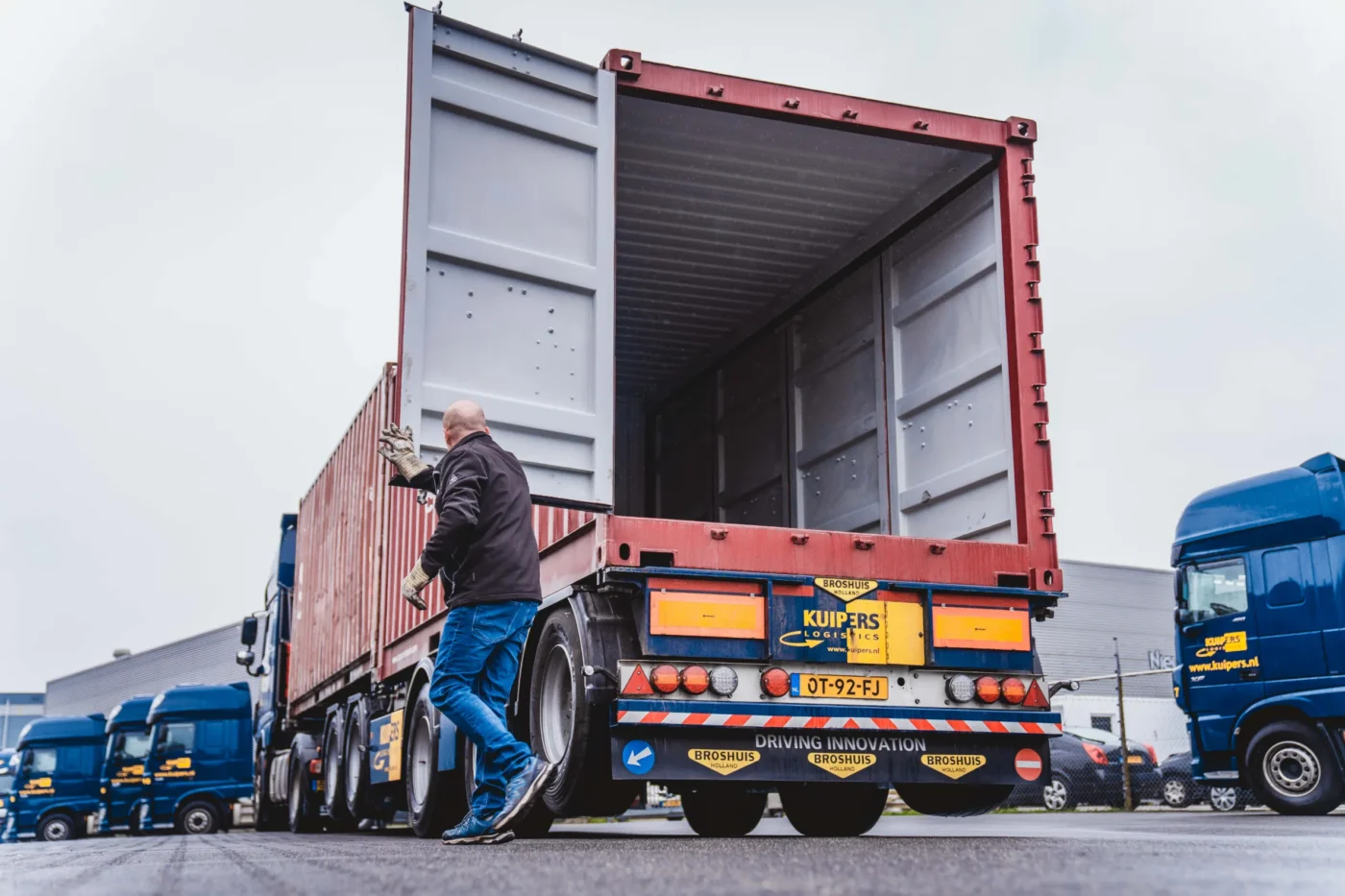 Een rode container wordt geopend. De container staat op een containerchassis van Kuipers Logistics en op de achtergrond staan de blauwe vrachtwagens van Kuipers Logstics op de parking.