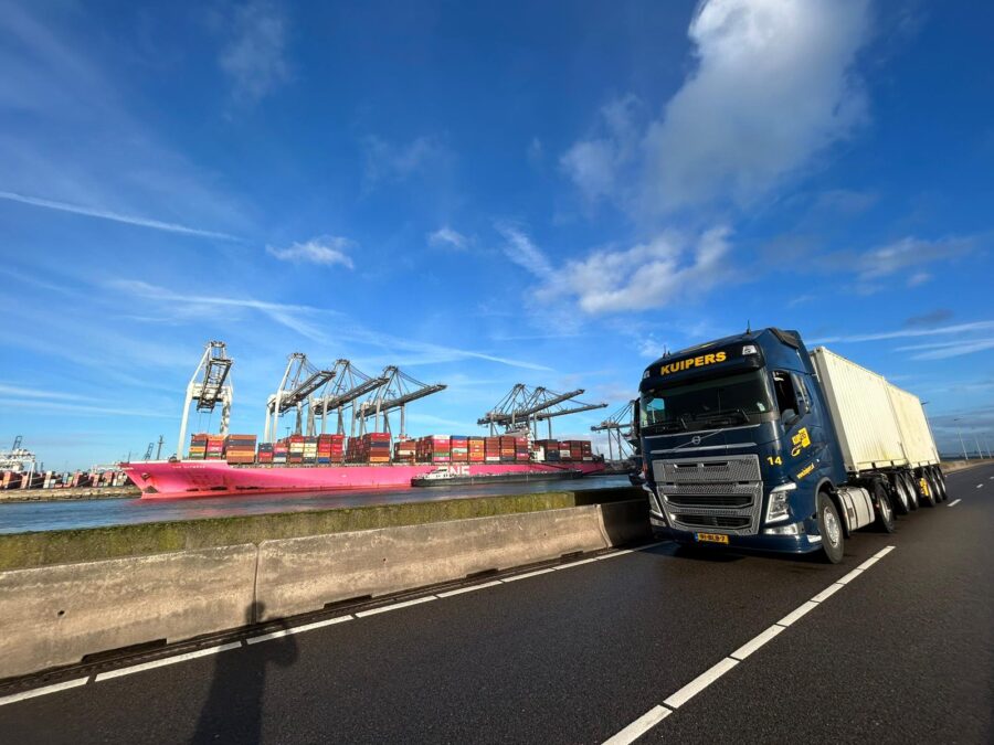 Een blauw met gele containervrachtwagen rijdt langs een internationaal containerschip. Op de vrachtwagen staan twee witte containers die perfect zijn voor internationaal transport.