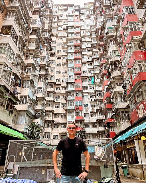 Wouter Engbersen staat voor een hoge flat in hongkong. Hij heeft een zwart tshirt aan en zondebril op en staat met zijn handen in zijn zij. Onder in de flat zitten winkeltjes.