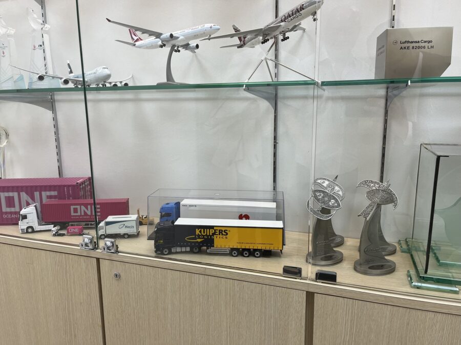 Een Kuipers Logistics vrachtwagen miniatuur staat prominent in de vitrine. Er omheen staan minaturen van vliegtuigen en vrachtwagens met containers. Ook staan er wat prijzen. De kuipers Logistics vrachtwagen is blauw met gele opdruk. 