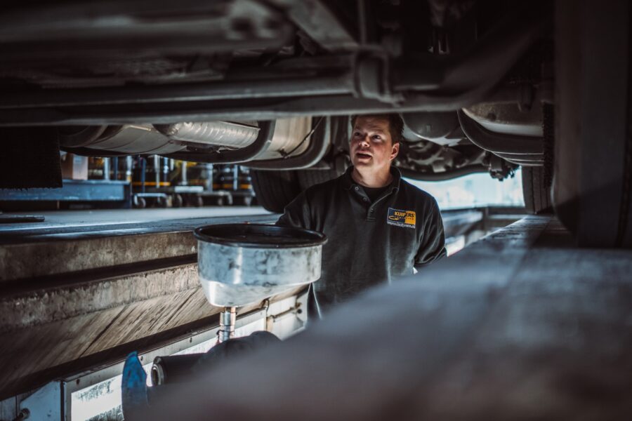 Wouter staat in de smeerkuil onder de vrachtwagen. Hij kijkt naar een onderdeel. Voor heb staat een ronde bak waar hij dingen in kan leggen.