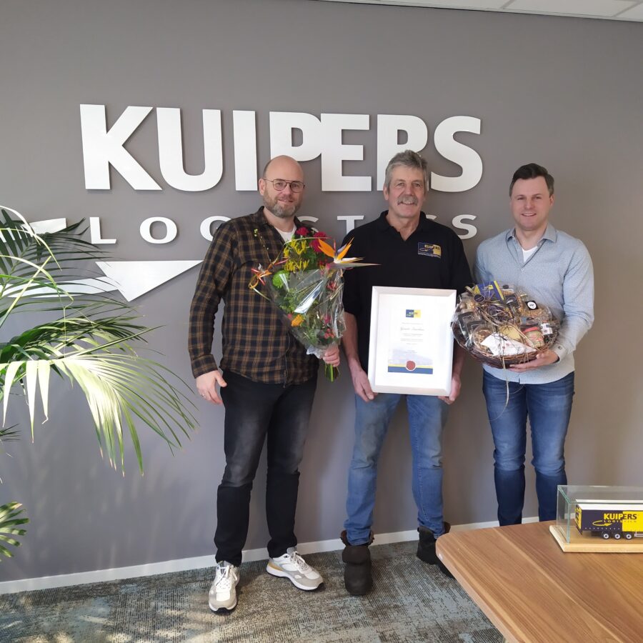 Gerard Laarhuis staat met zijn oorkonde tussen Philip Vollenbroek en Thijs Lansink voor het Kuipers Logistics logo. Philip heeft een bos bloemen vast en Thijs een gevulde mand met allemaal eten en drinken.
