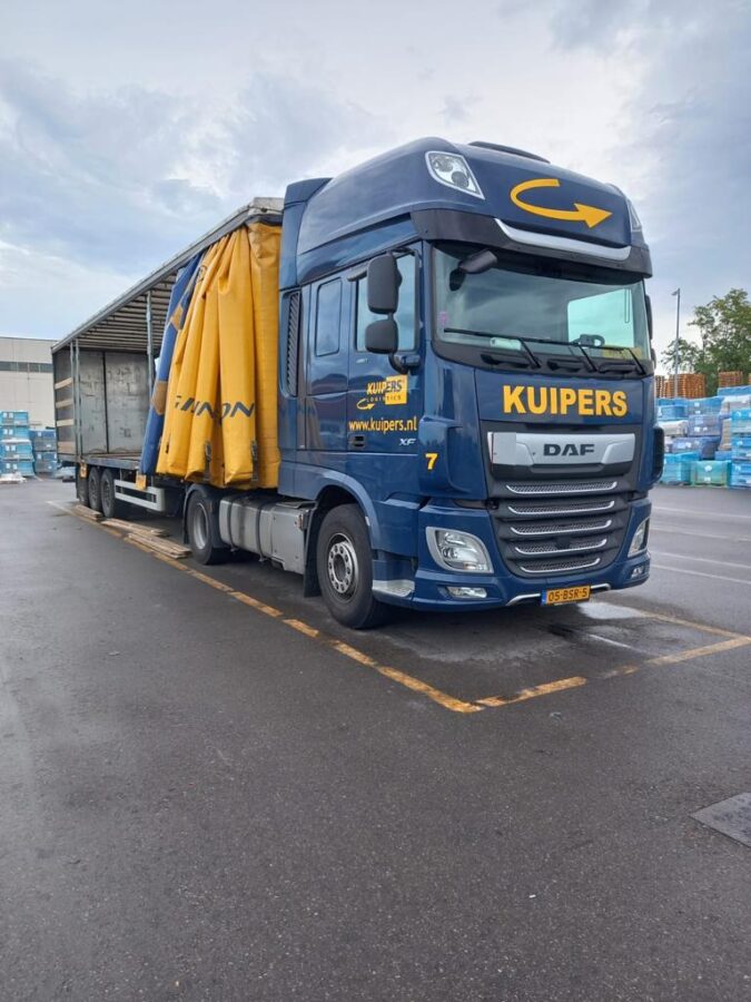 Een blauw daf xf 480 van Kuipers Logistics staan in een geel omlijnd parkeervak. Om de vrachtwagen heen staan allemaal tegels die in blauw folie zijn gewikkeld. De Zeilen van de trailer staan open en er zijn al wat planken uitgehaald, zodat er makkelijk geladen kan worden.