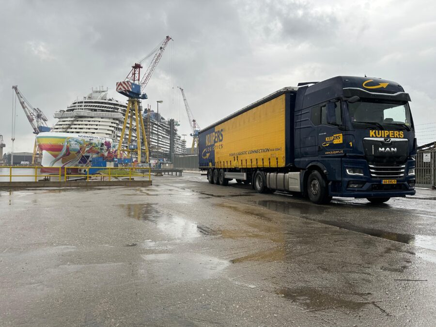 Een MAN TGX 480 van Kuipers Logistics staat in de Haven van Venetië op een regenachtige dag. De Wolken zijn grauw en de weg nat. Waardoor de blauw met gele vrachtwagen goed opvalt. De vrachtwagen staat voor een groot cruiseschip. De Norwegian Aqua. Een boot waar en in totaal 3 van zijn gemaakt. op de voorkant zijn vele kleurrijke strepen te zien. Het is nog net geen regenboogvlag. 