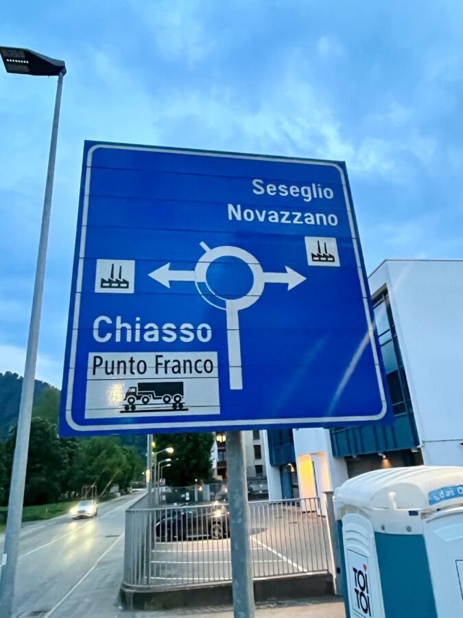 Een blauw verkeersbord van een rotonde. Links ga je naar het industrieterrein van Chiasso en rechs ga je naar Seseglio of Novazzano.