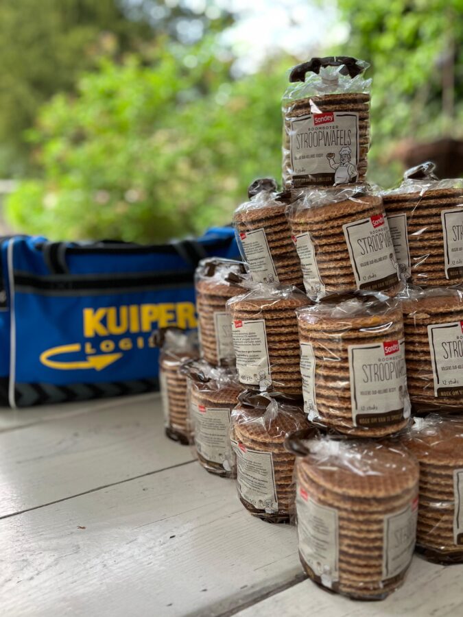 Stroopwafels van het merk Sodney staan in een piramide gestapeld op een tuintafel. Achter de stroopwafels is een Kuipers Logistics tas zichtbaar. En wazig is het groen van de tuin nog te zien.