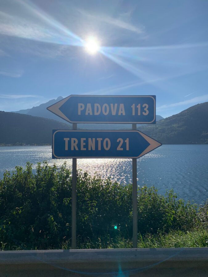 Wegwijzing aan het water. Links gaat naar Padova. Rechts naar Trento. De zon schijnt op de achtergrond.