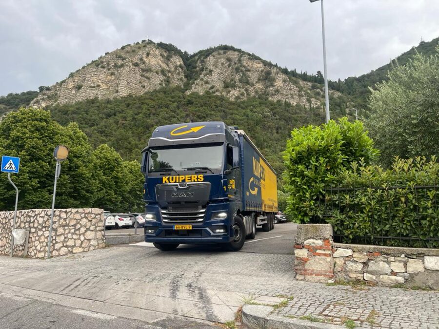 Een MAN TGX 18.480 rijdt van de parkeerplaats. De MAN is Blauw en is bedrukt met gele logo's van Kuipers Logistics. Achter de vrachtwagen zit ook een trailer van Kuipers Logistics. Die is half blauw half geel. Ook de trailer heeft groot het logo. Het logo wordt gesplits door het blauw gele en is geel op het blauwe deel van de trailer en blauw op het gele deel van de trailer.