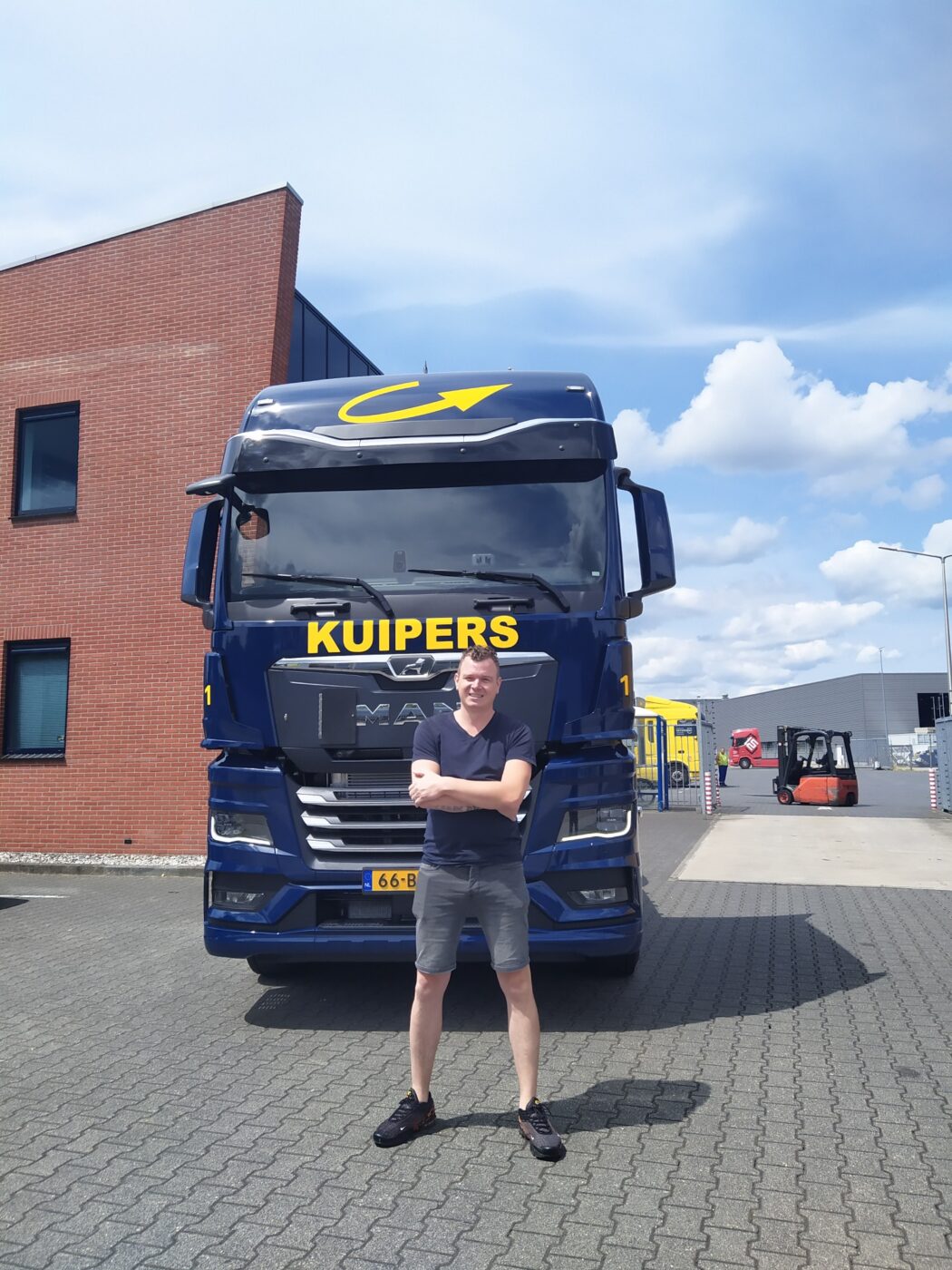 Patrick staat voor de MAN TGX 18.480. hij lacht, maar kijk wel moeilijk, omdat hij tegen de zon in kijkt. Hij draag een t-shit met korte broek.