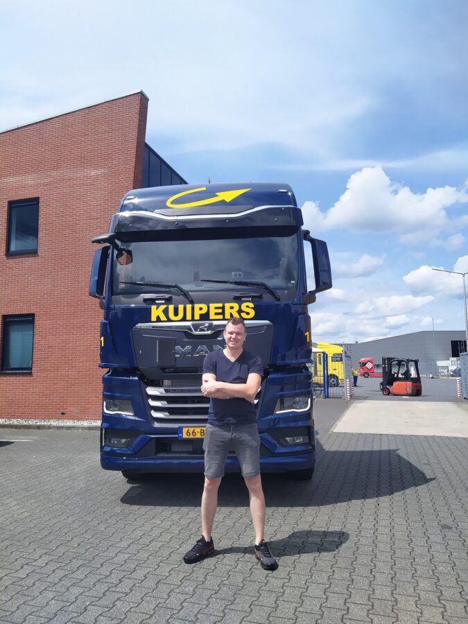 Patrick staat voor de MAN TGX 18.480. hij lacht, maar kijk wel moeilijk, omdat hij tegen de zon in kijkt. Hij draag een t-shit met korte broek. 