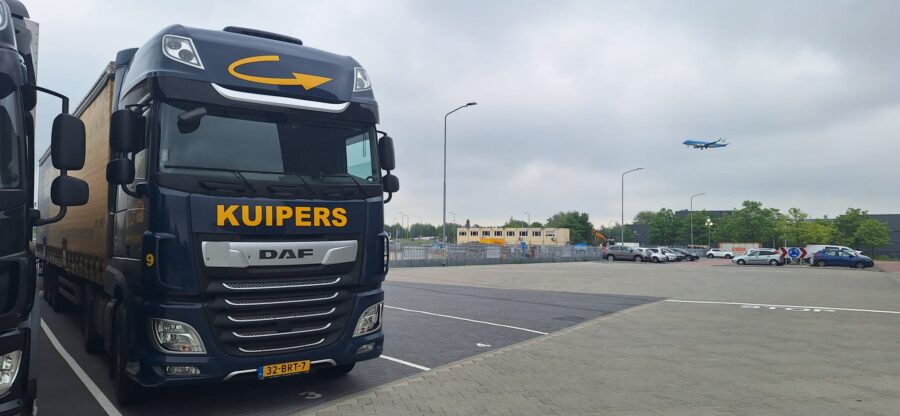 Een blauw met gele vrachtwagen staat op de parkeerplaats. Achter de vrachtwagen in de lucht is een vliegtuig te zien.