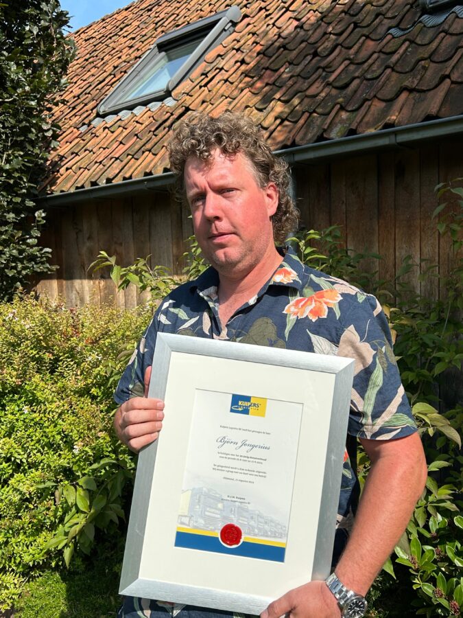 Bjorn Jongerius staat op de foto met zijn oorkonde. Hij staat in zijn tuin en draagt een blouseje met bloemen er op. De zon schijnt in zijn gezicht.
