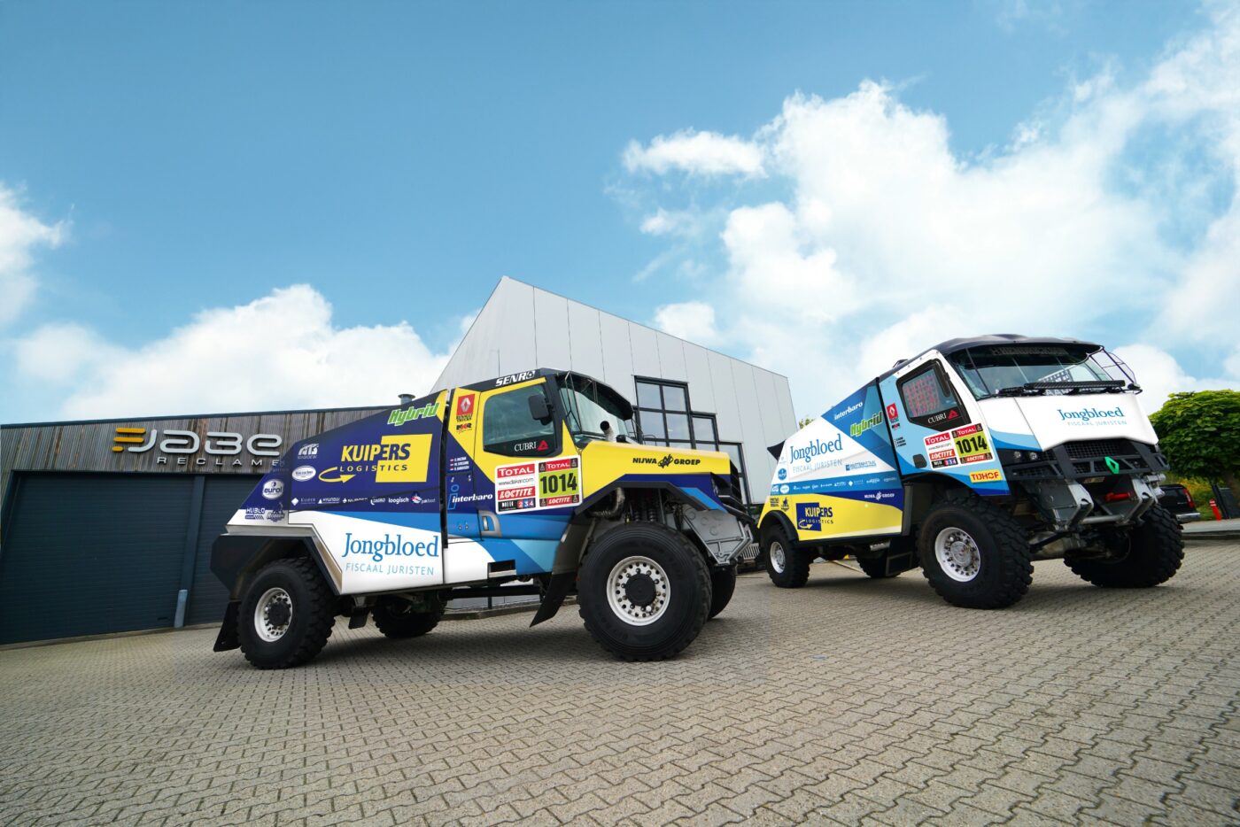alt="Twee rally vrachtwagens staan op een plas. Op de vrachtwagens is de bedrukking Kuipers Logistics en Jongbloed Fiscaal Juristen groot zichtbaar. De ene truck meer in de blauw met gele kleuren van Kuipers Logistics. De andere truck meer in de wit met blauwe kleuren van Jongbloed fiscaal juristen. "