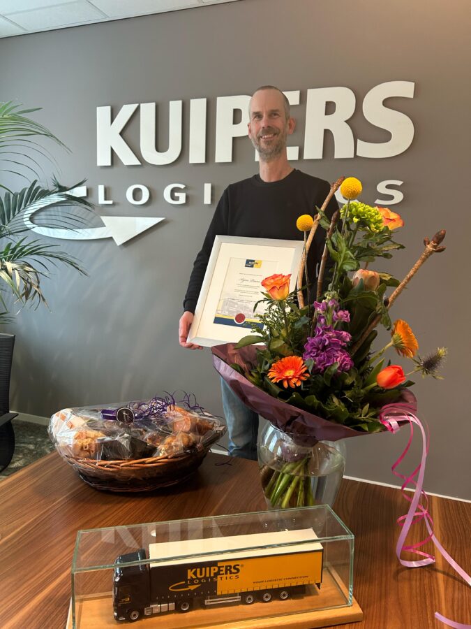 Arjan Paans staat in de vergaderzaal van Kuipers Logistics voor het bedrijfslogo. Op de tafel staat een geschenkmand, een bos bloemen en een miniatuur van een Kuipers Logistics truck. In zijn hand heeft hij de oorkonde die hij heeft gekregen voor zijn 12.5 jarige dienstverband.