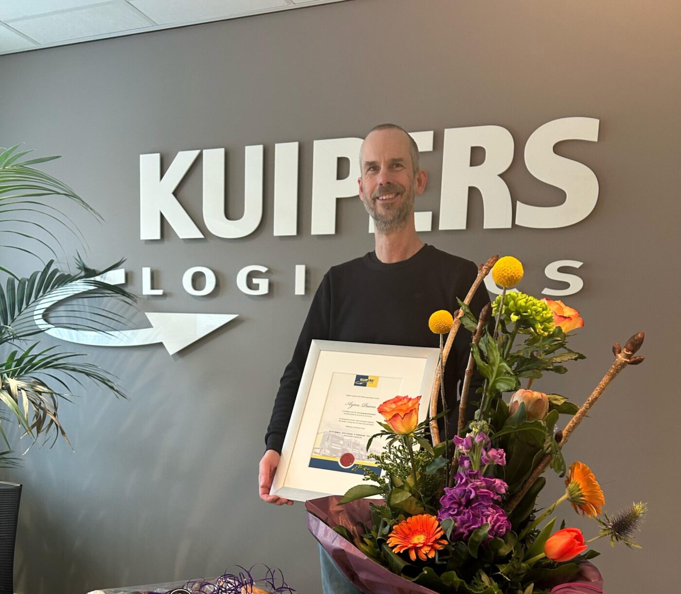 Arjan Paans staat in de vergaderzaal van Kuipers Logistics voor het bedrijfslogo. Op de tafel staat een geschenkmand, een bos bloemen en een miniatuur van een Kuipers Logistics truck. In zijn hand heeft hij de oorkonde die hij heeft gekregen voor zijn 12.5 jarige dienstverband.