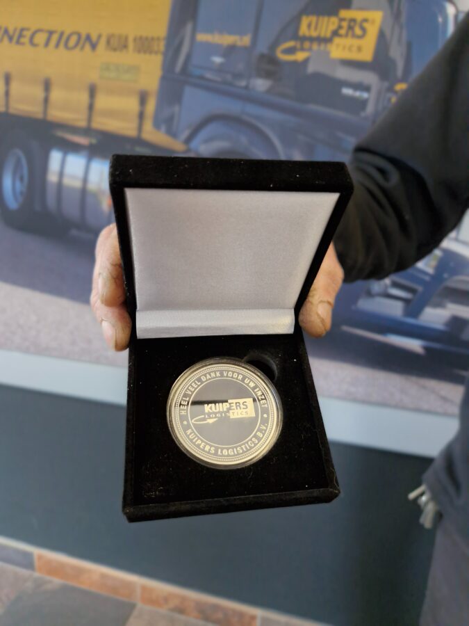 De zilveren Kuipers Logistics penning met witte opdruk. Op de penning staat de tekst:: "heel veel dank voor uw inzet Kuipers Logistics B.V. " iIn het midden staat het Kuipers Logistics logo