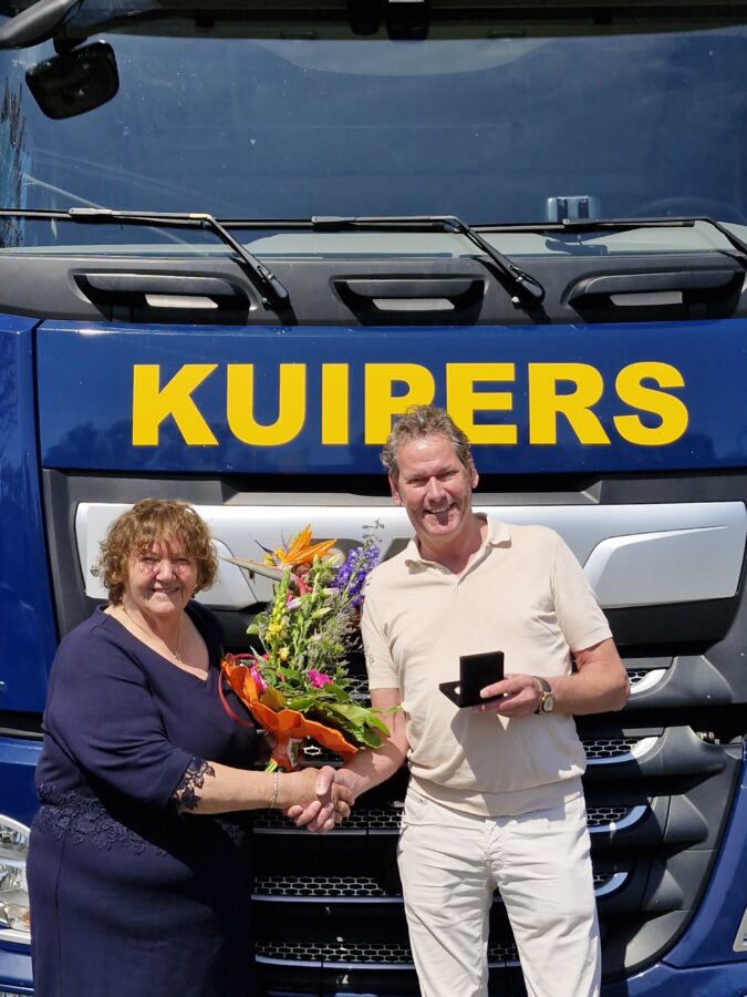 Maria en Harry staan voor de Kuipers truck waar Maria in gereden heeft. Maria heeft bloemen vast in haar linker hand. Met rechts schud ze de hand van Harry. Harry houd in zijn andere hand de ere penning vast