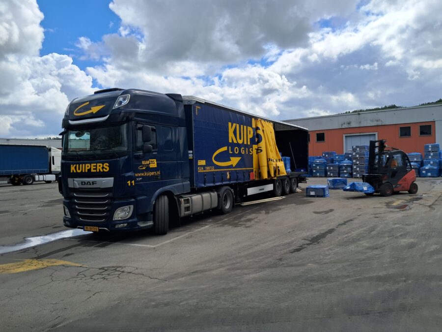 Een heftruck laad tegels in de Kuipers Logistics vrachtwagen via de zijkant van de vrachtwagen. De tegels zijn gewikkeld in blauw plastic en er staan er achter de vrachtwagen nog veel meer. 